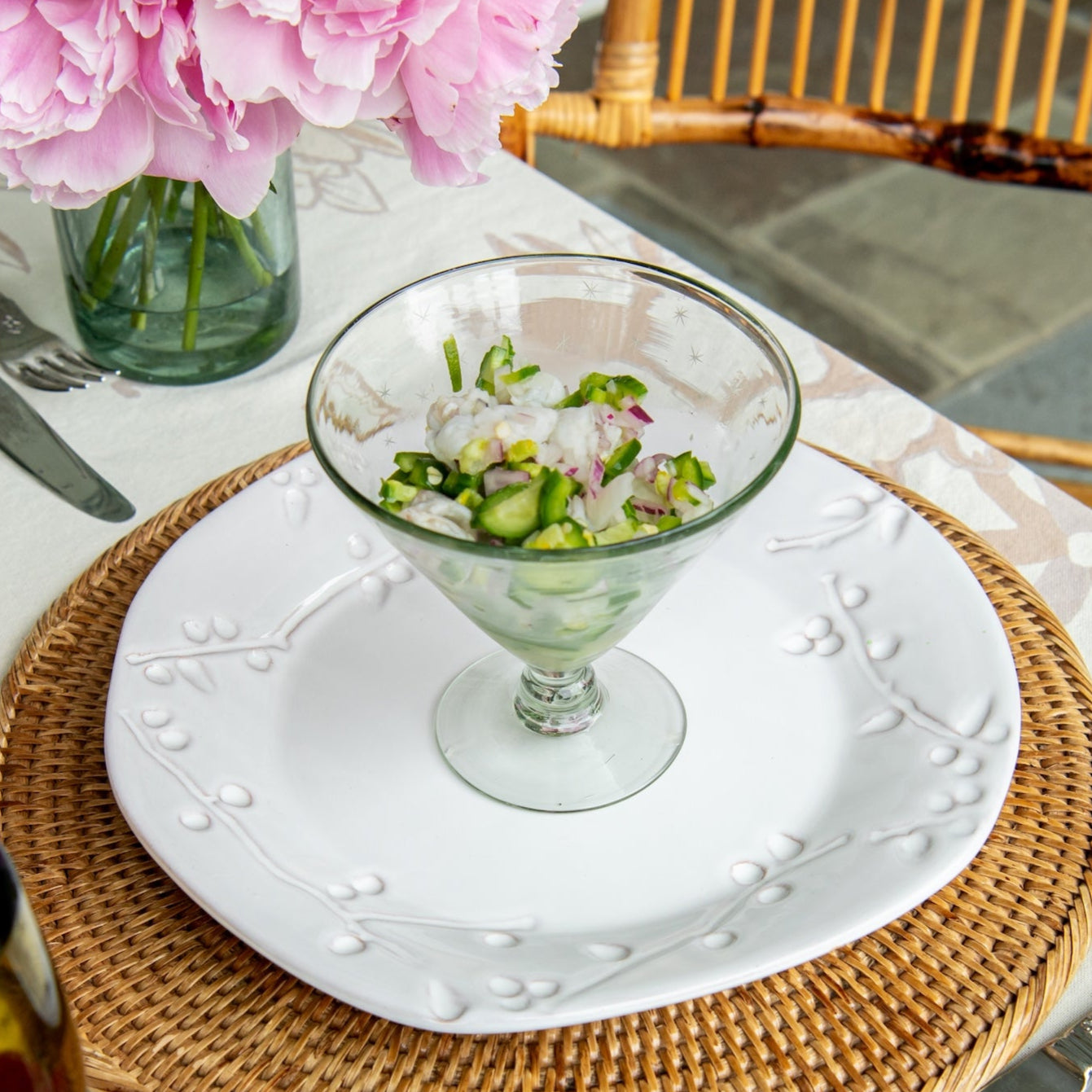 Dining table setting with pink flowers, glass dish, and cutlery on a white tablecloth.