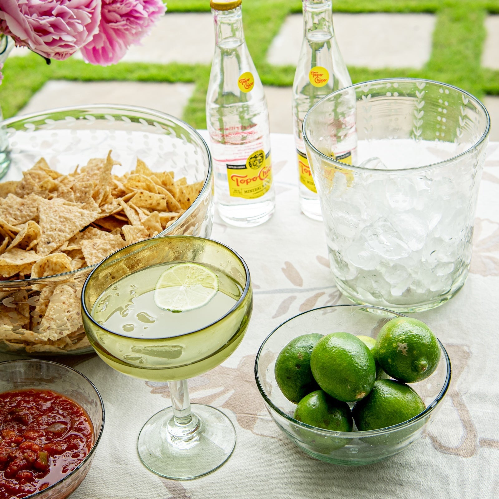 Outdoor setting with tortilla chips on a clear etched bowl, limes in a small etched bowl, a glass of limeade, and bottles on a table.
