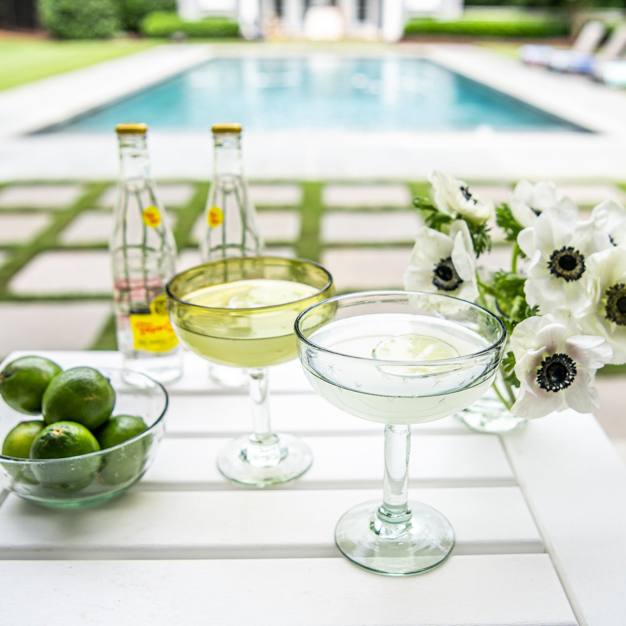 Outdoor setting with margarita glasses filled with cocktails, limes, and flowers by a poolside.