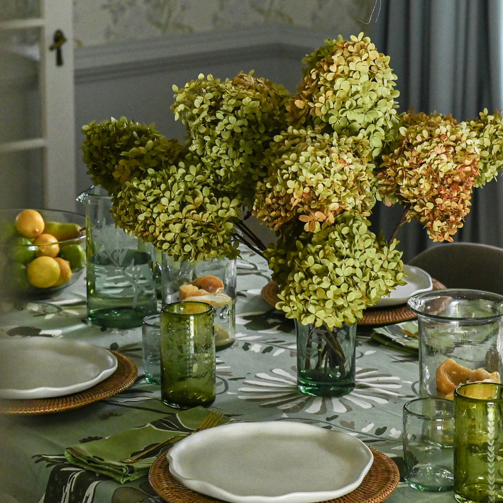 Dining table set with floral centerpiece, plates, and glasses in a room with curtains and a window.