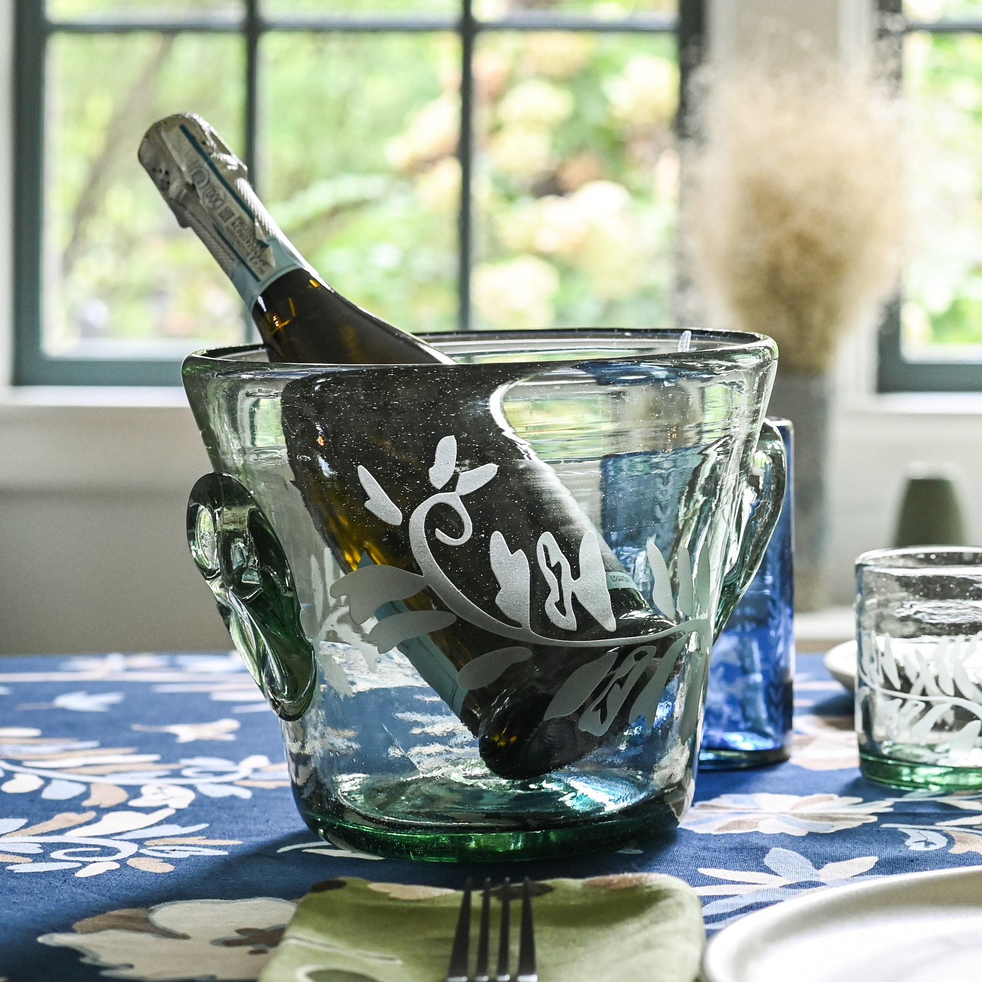 A clear glass champagne bucket with hand-etched fern frond motif, containing a champagne bottle, placed on a table with a blue and white floral tablecloth.