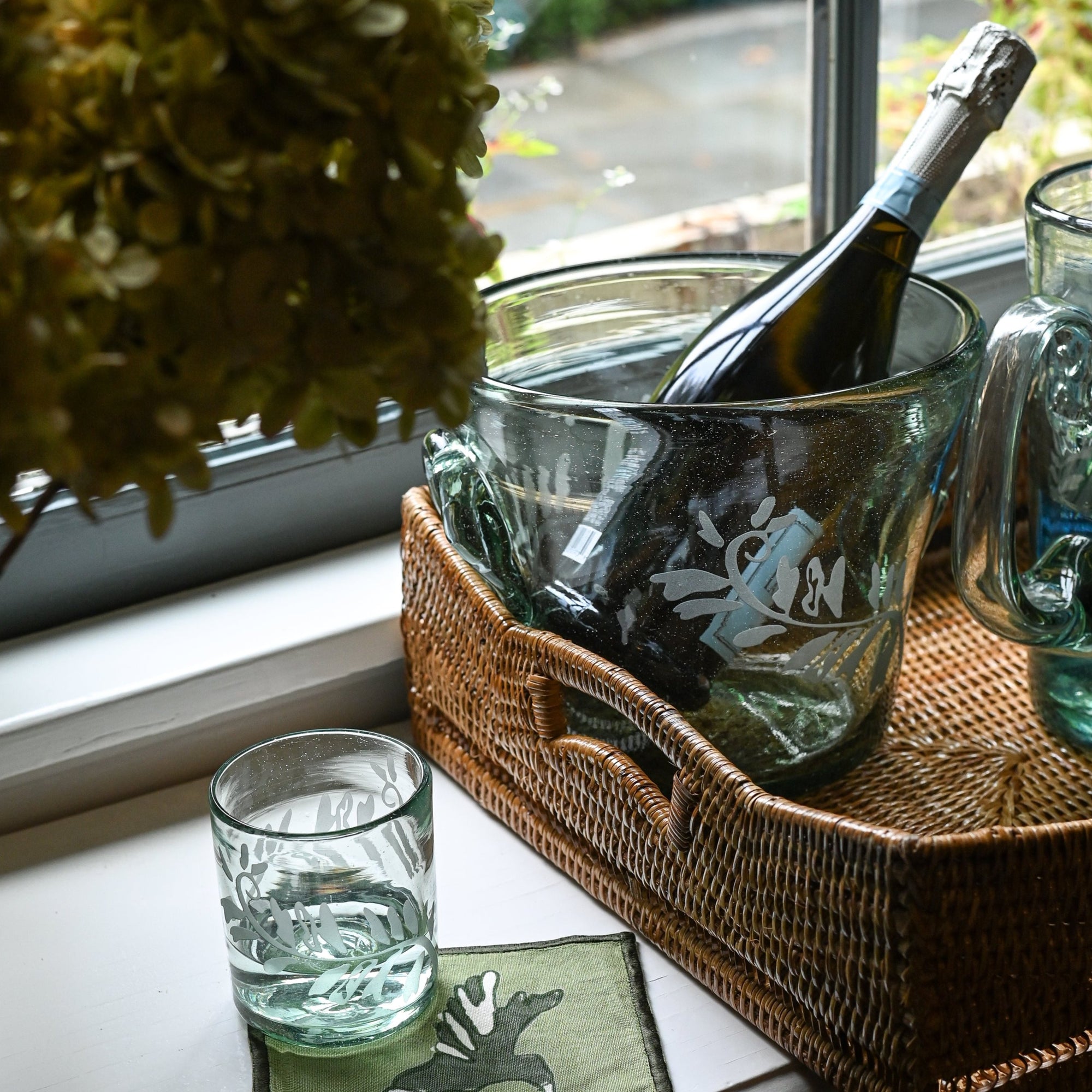 A clear glass champagne bucket with hand-etched fern frond motif, containing a champagne bottle, placed on a table with a blue and white floral tablecloth.