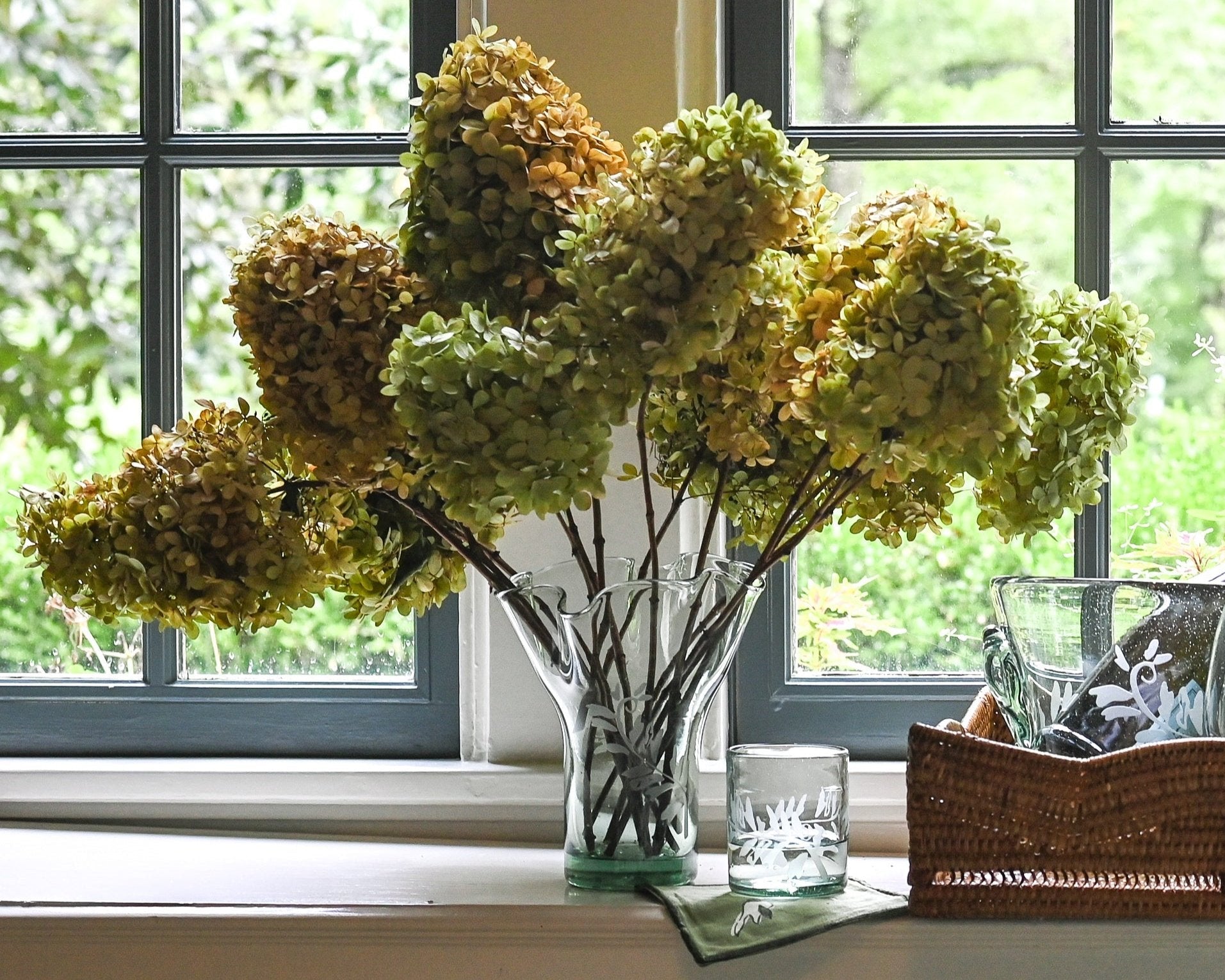 Bouquet of hydrangeas in a vase on a windowsill with a glass and basket in the background.