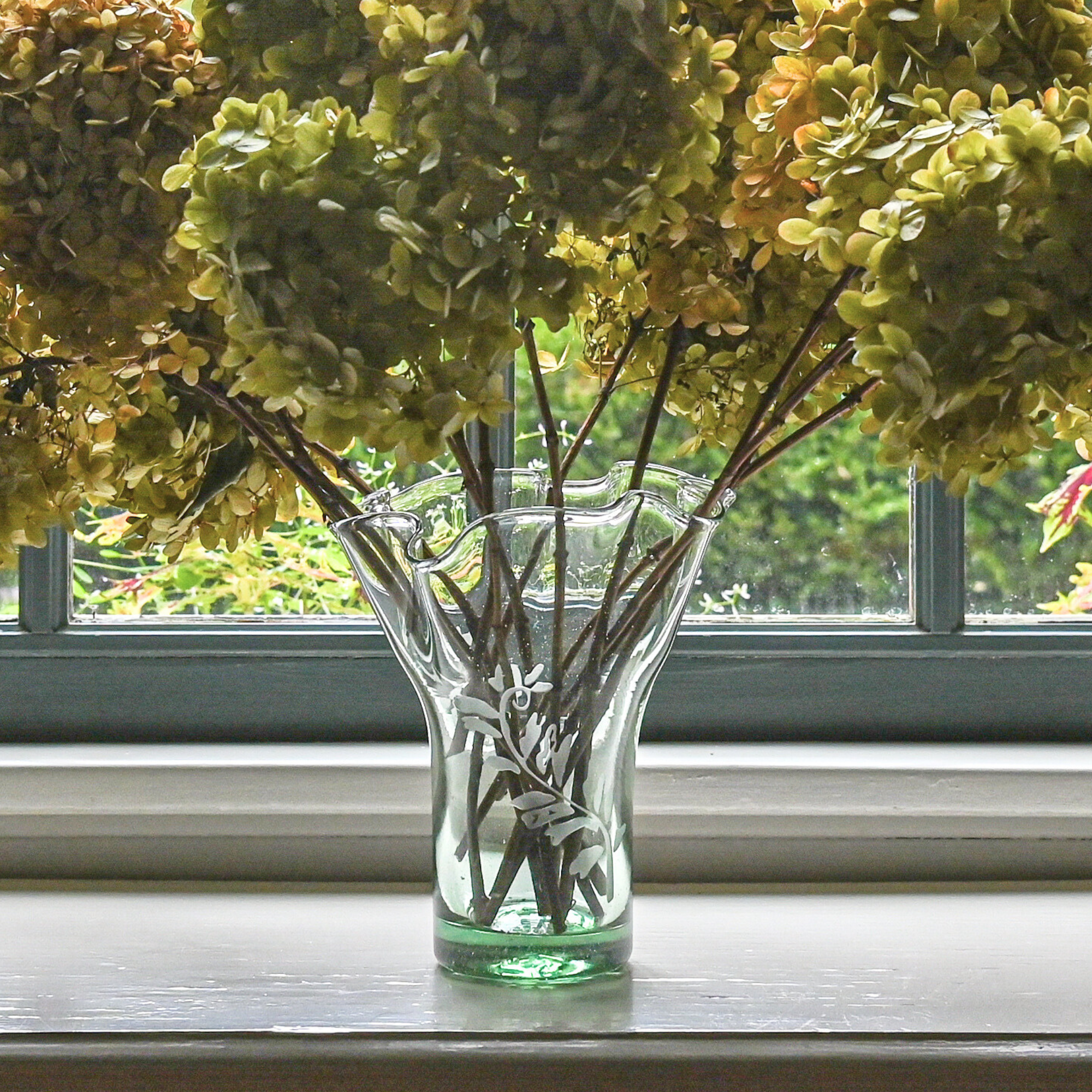 A clear glass vase with a fern leaf motif, containing stems and placed on a windowsill with hydrangea flowers in the background.