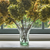 A clear glass vase with a fern leaf motif, containing stems and placed on a windowsill with hydrangea flowers in the background.