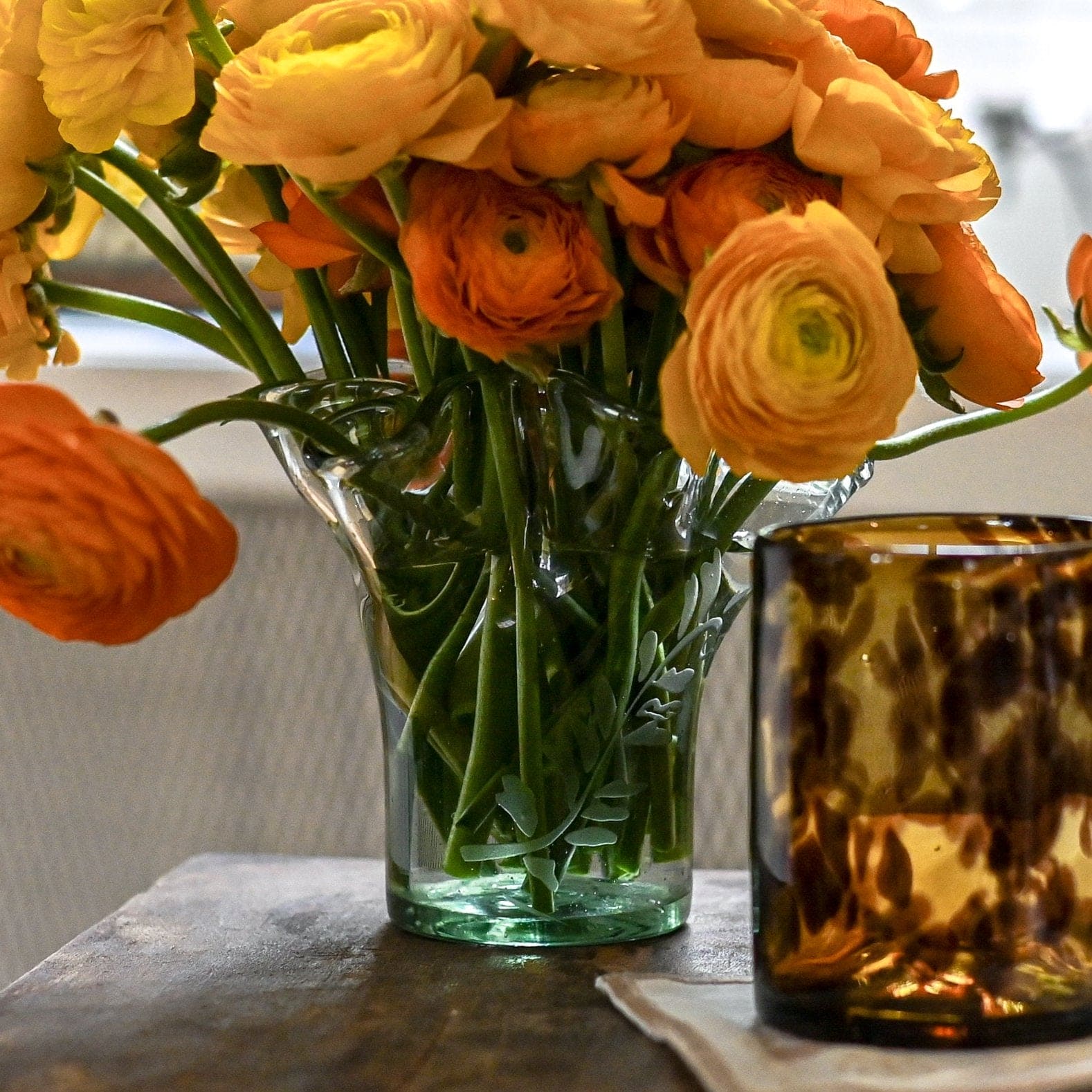 A clear glass vase with a fern motif, containing orange and yellow flowers, placed on a surface with a brown glass object beside it.