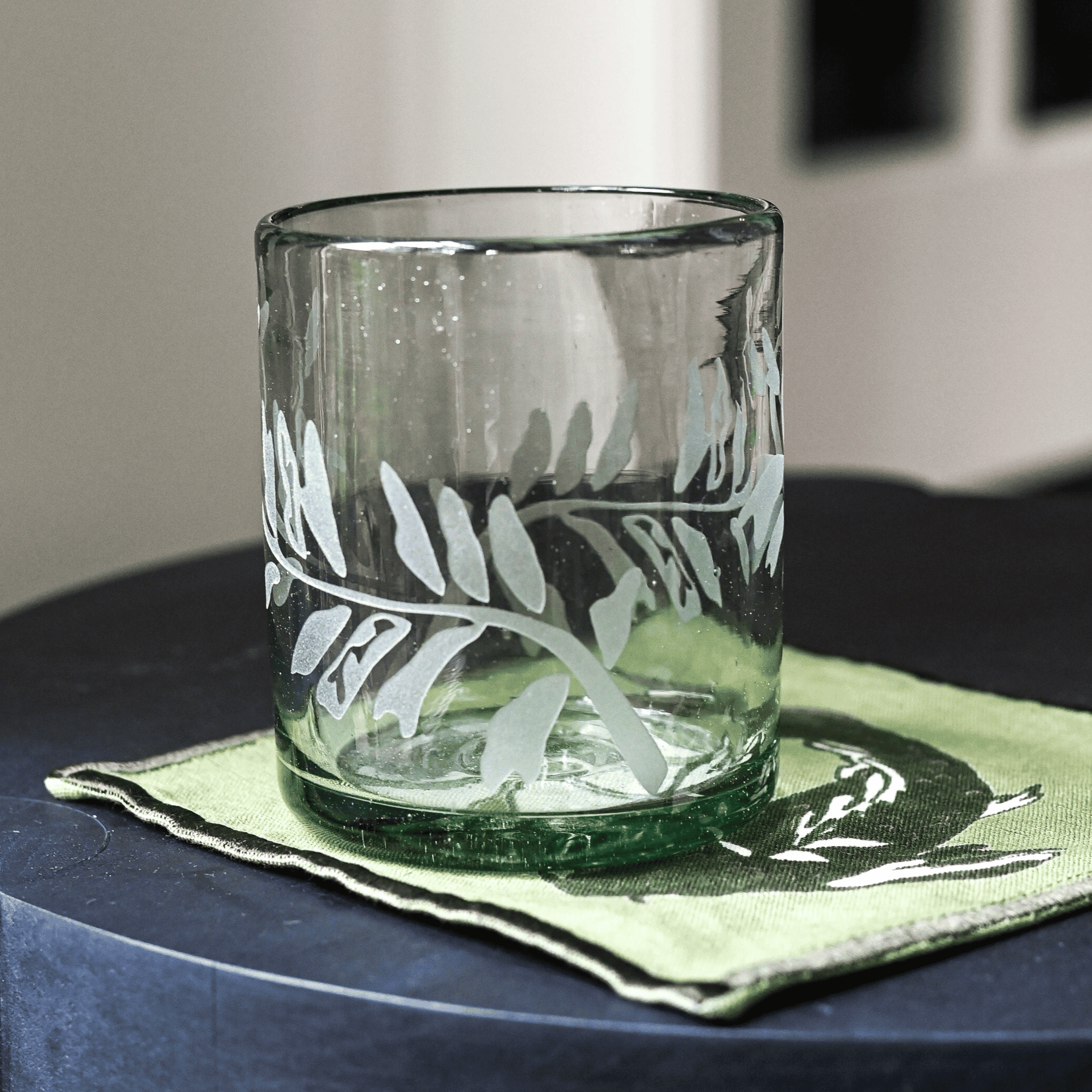 A clear rocks glass with hand-etched fern frond motif, placed on a green coaster on a dark surface.