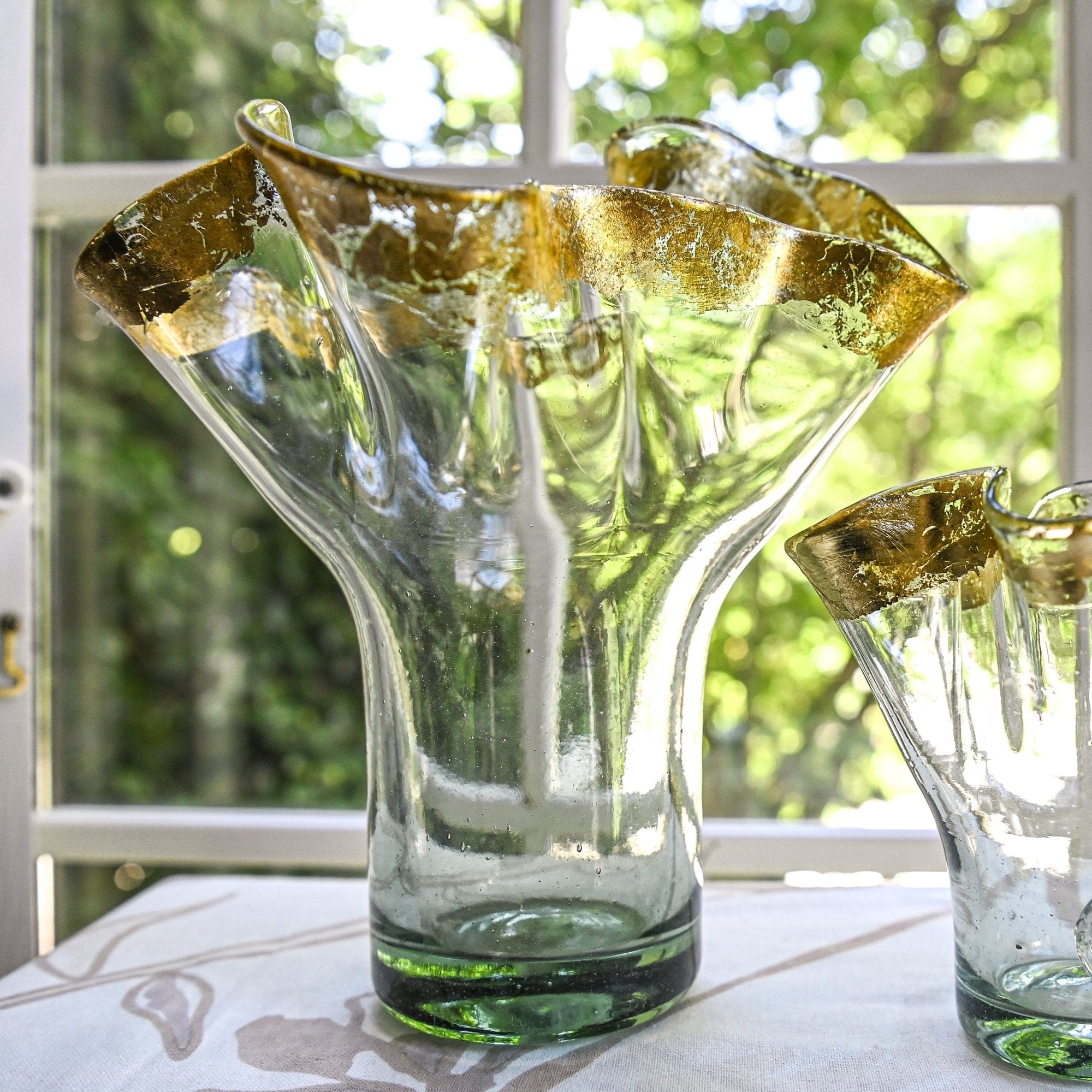 A clear glass vase with gold leaf ribbon rim, displayed in front of a window with a green outdoor background.