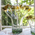 A clear glass vase with gold leaf ribbon rim, displayed in front of a window with a green outdoor background.
