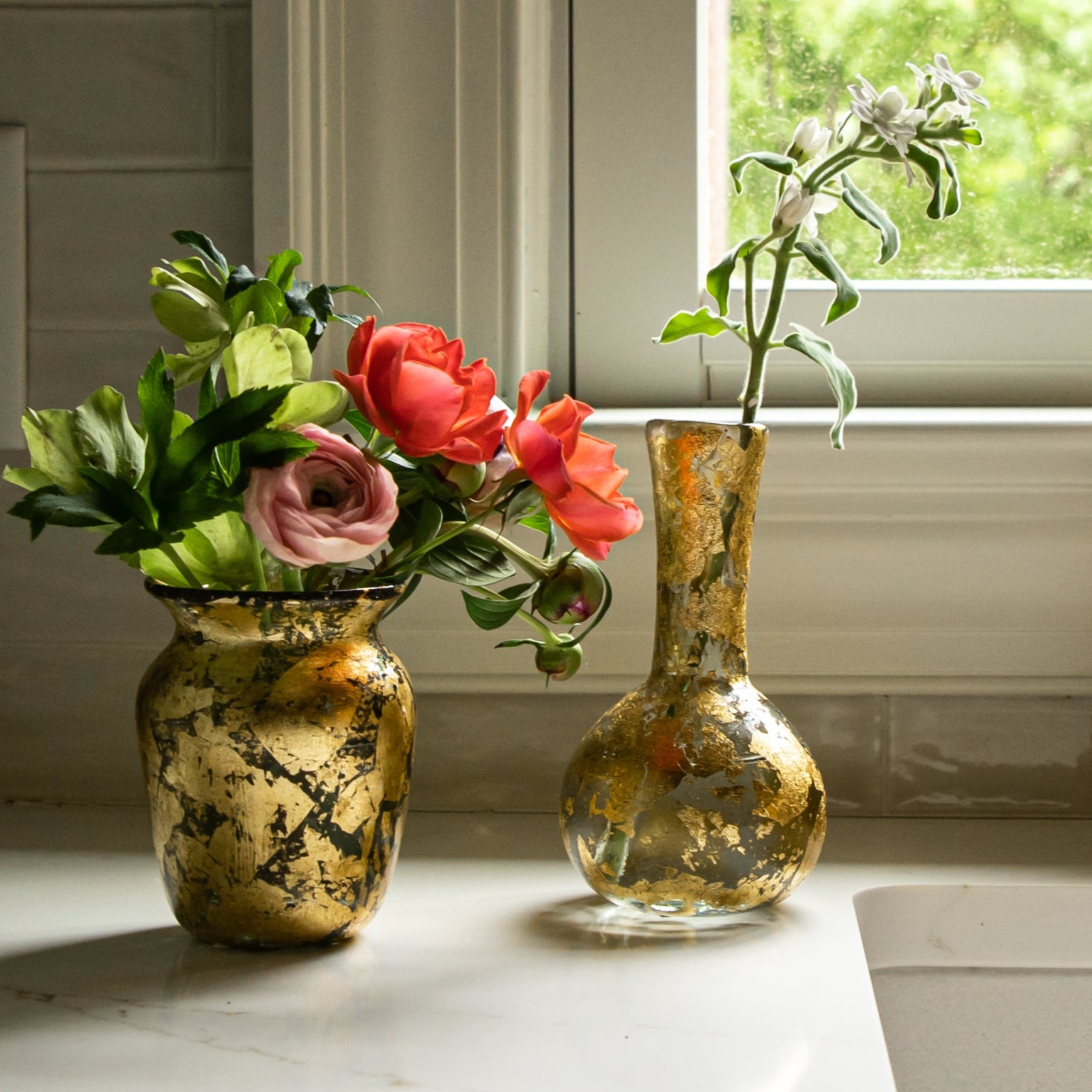 Kitchen sink with brass faucet, flowers in vases covered in 14k gold leaf, and window in the background