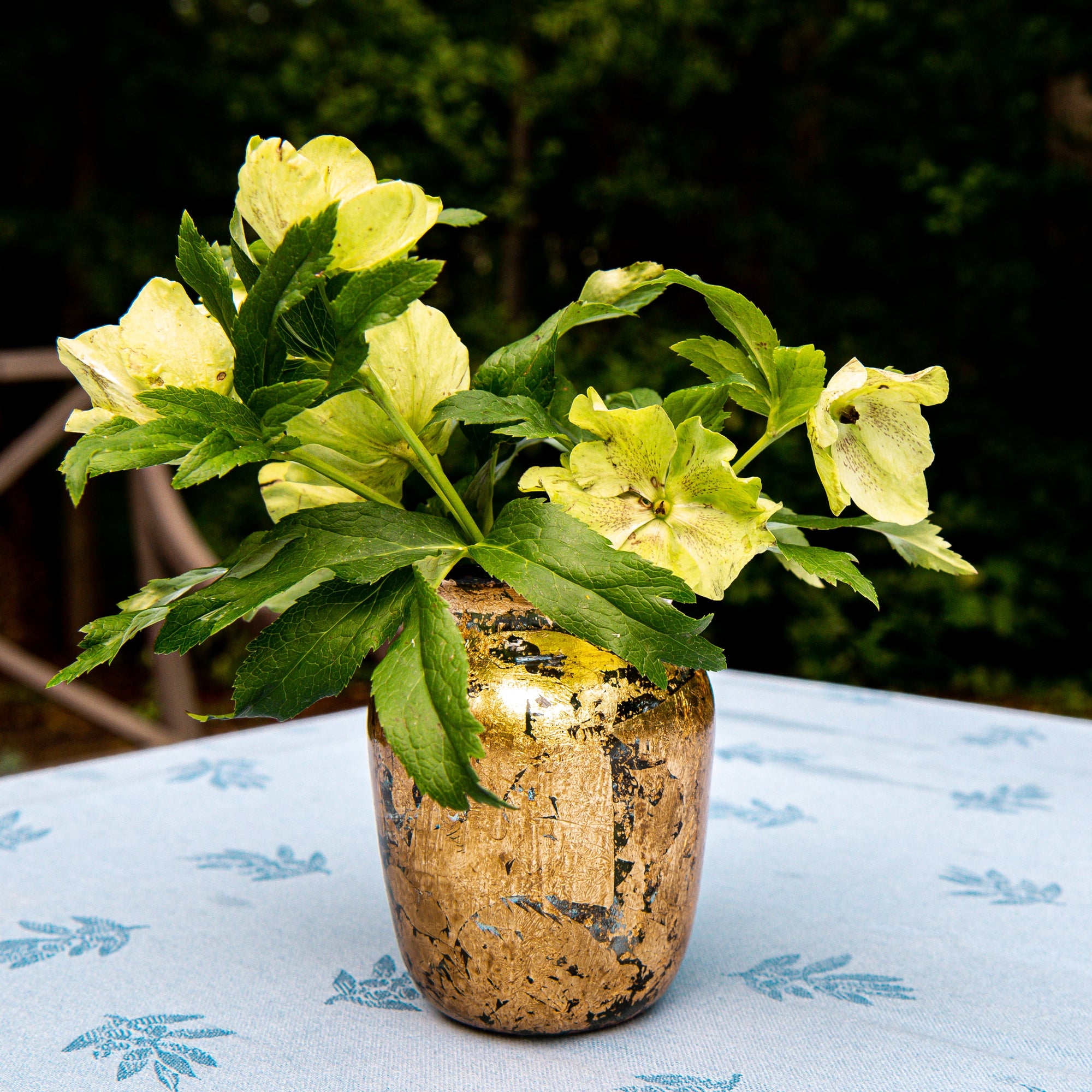 Gold vase with greenery and yellow flowers on a tablecloth outdoors
