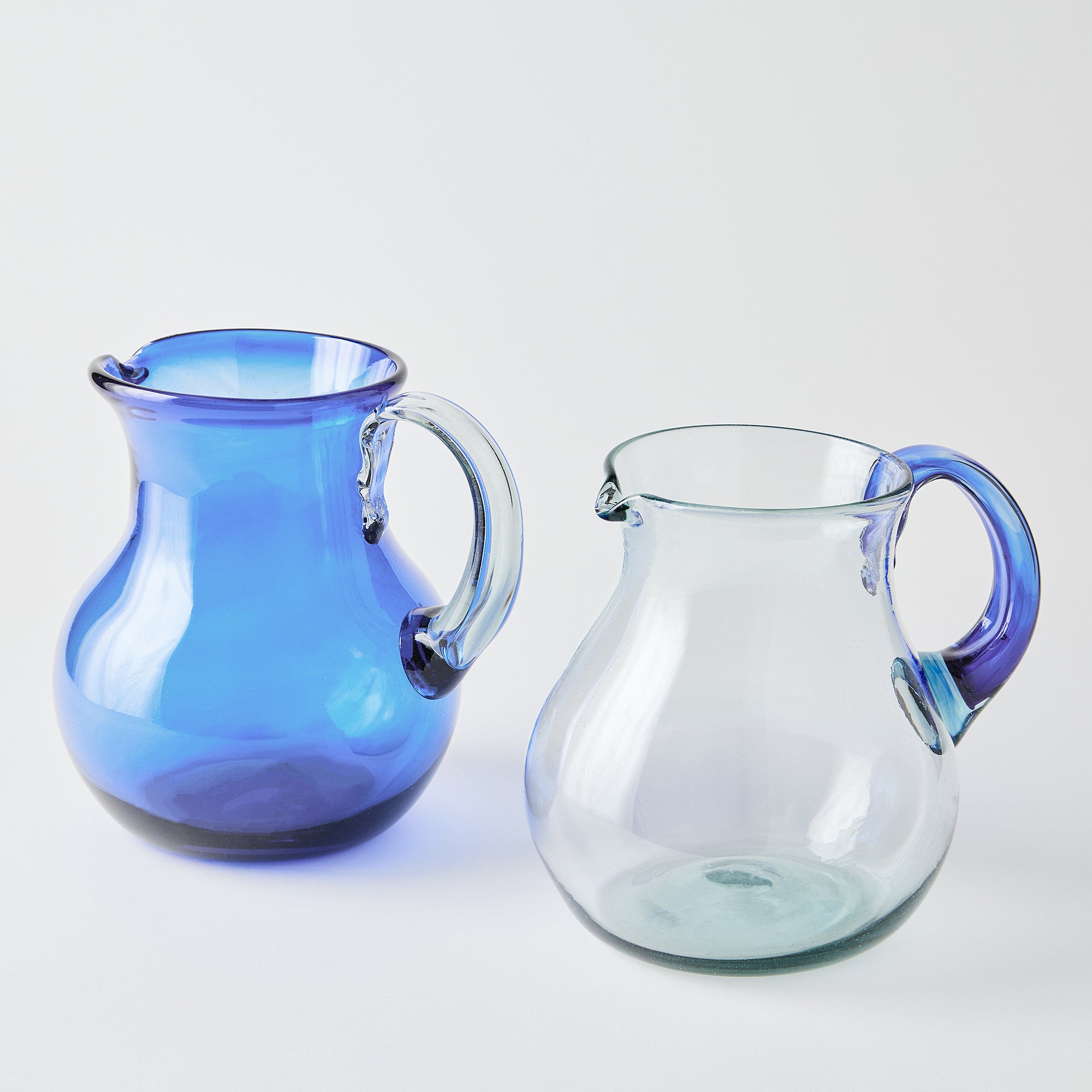 Two glass pitchers, one blue with a clear handle and one clear with a blue handle, on a white background