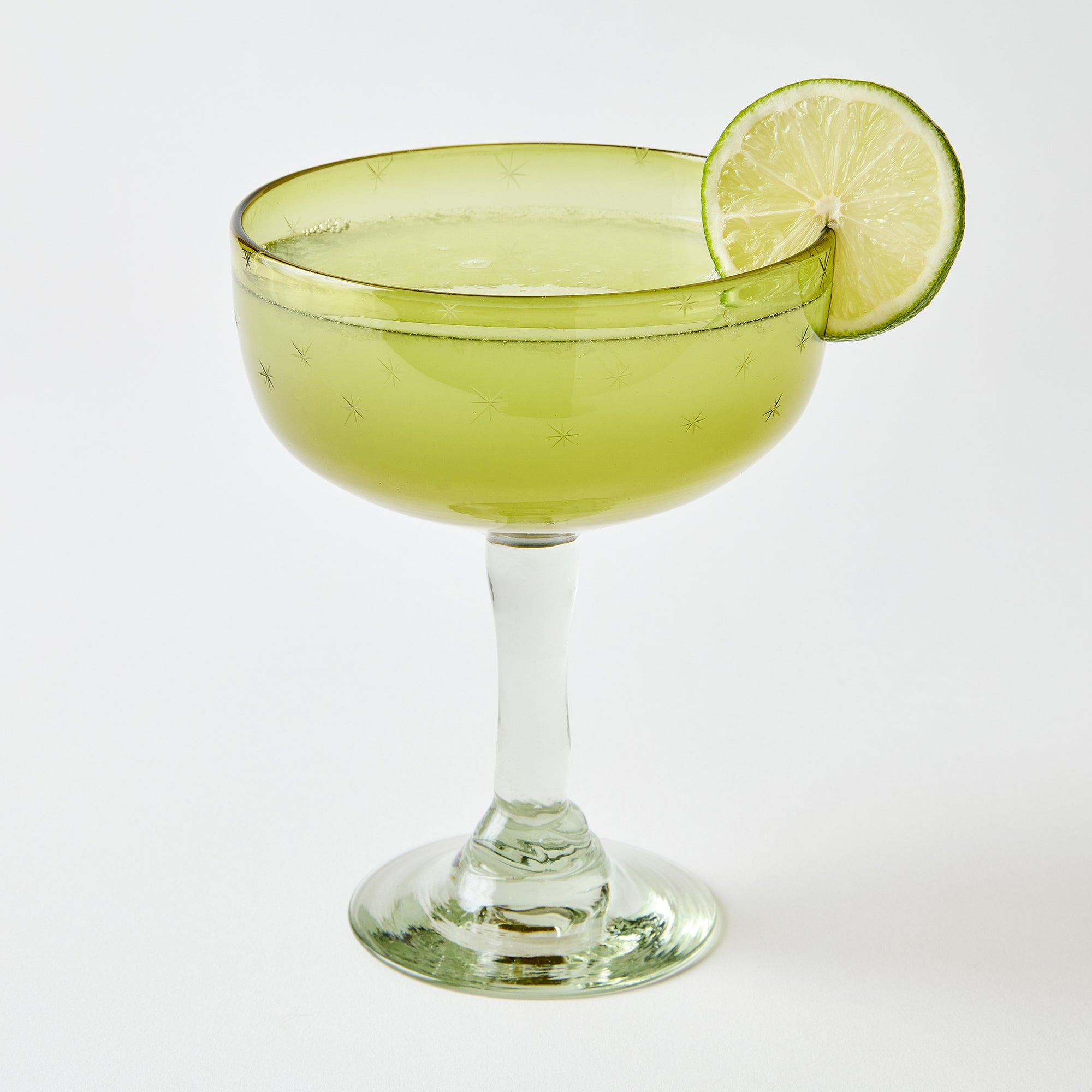 Cocktail in a yellow margarita glass etched with stars with a lime wheel on a white background