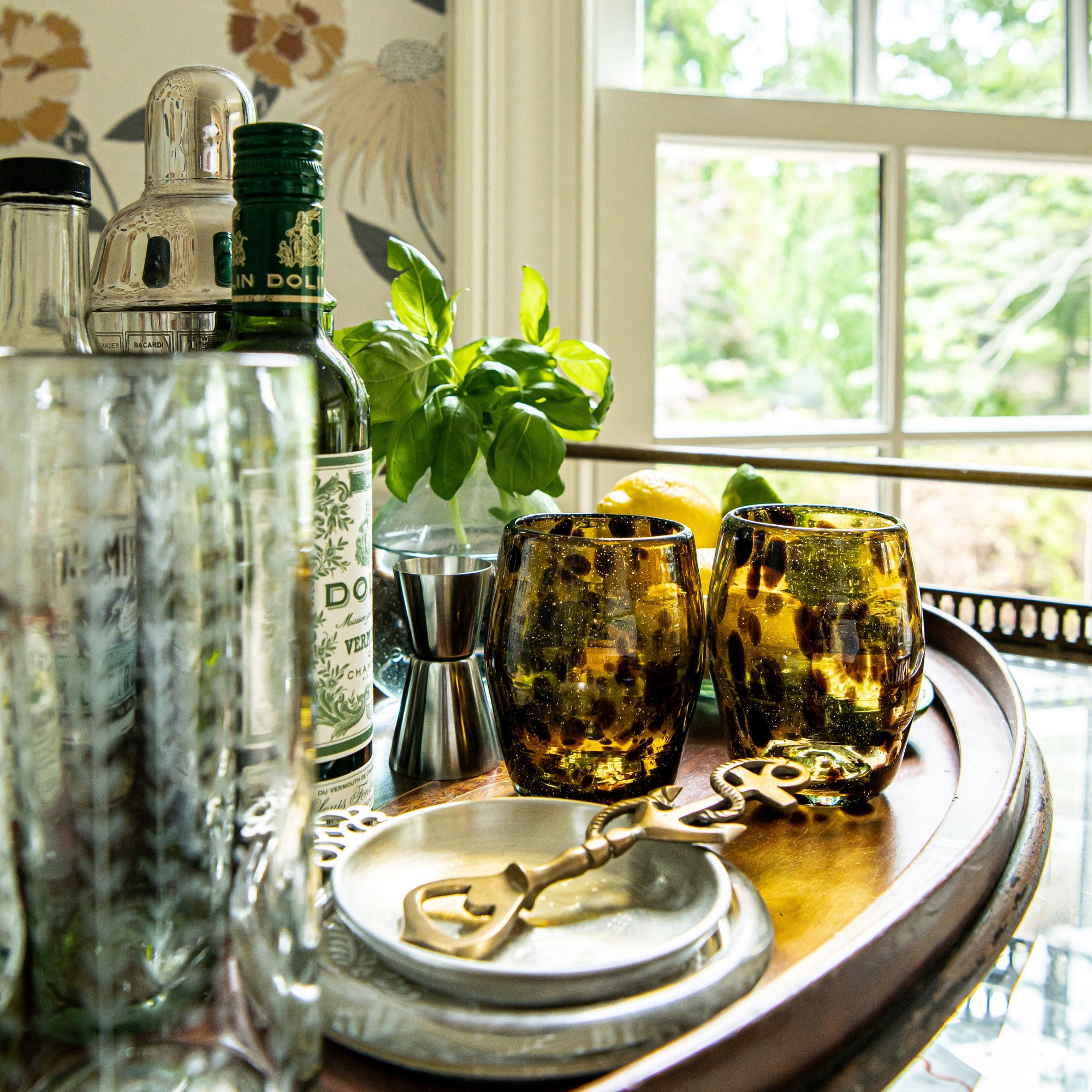 Alcohol bottles, glasses, and bar tools on a wooden surface with a window in the background