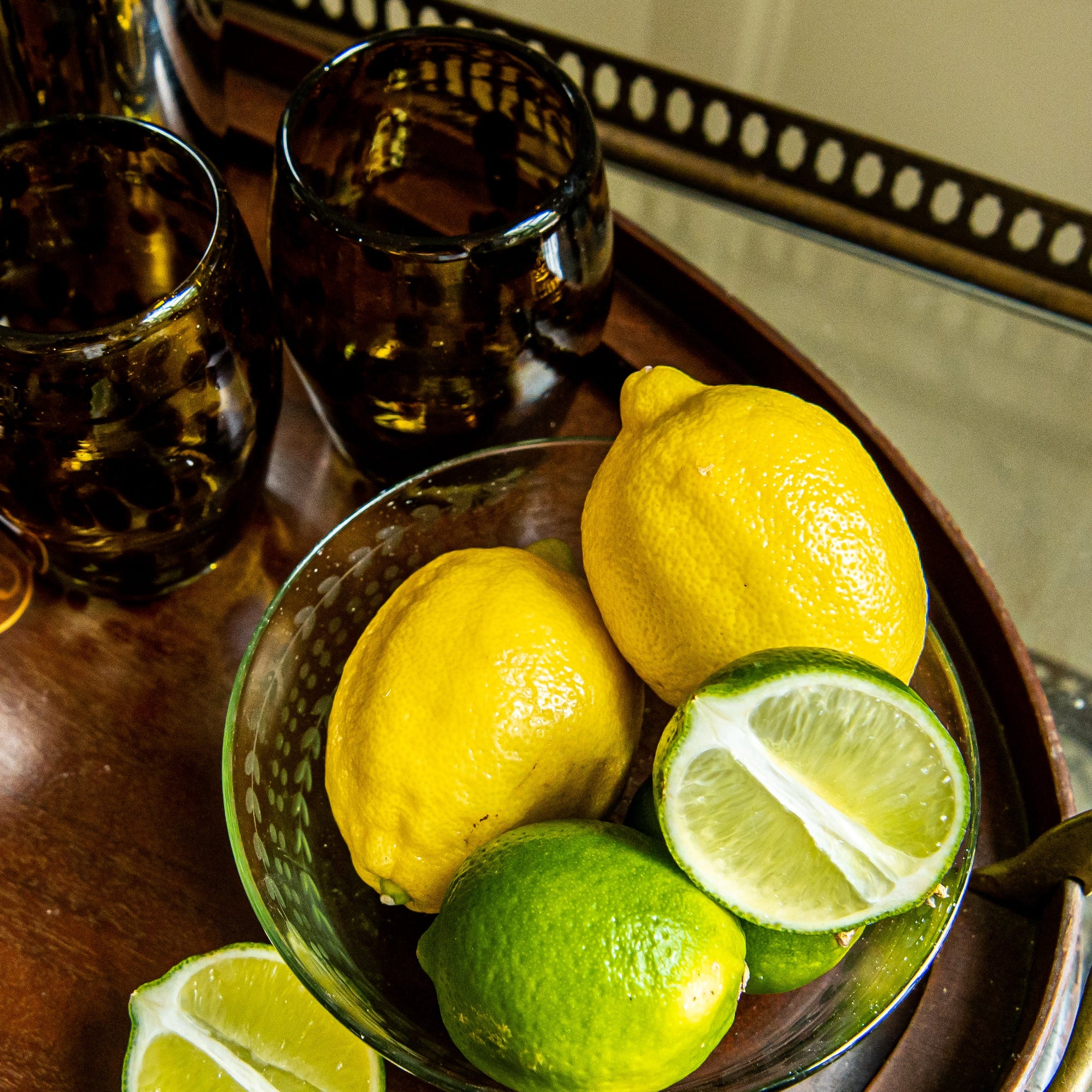 Bowl of lemons and limes on a wooden tray with glasses in the background.