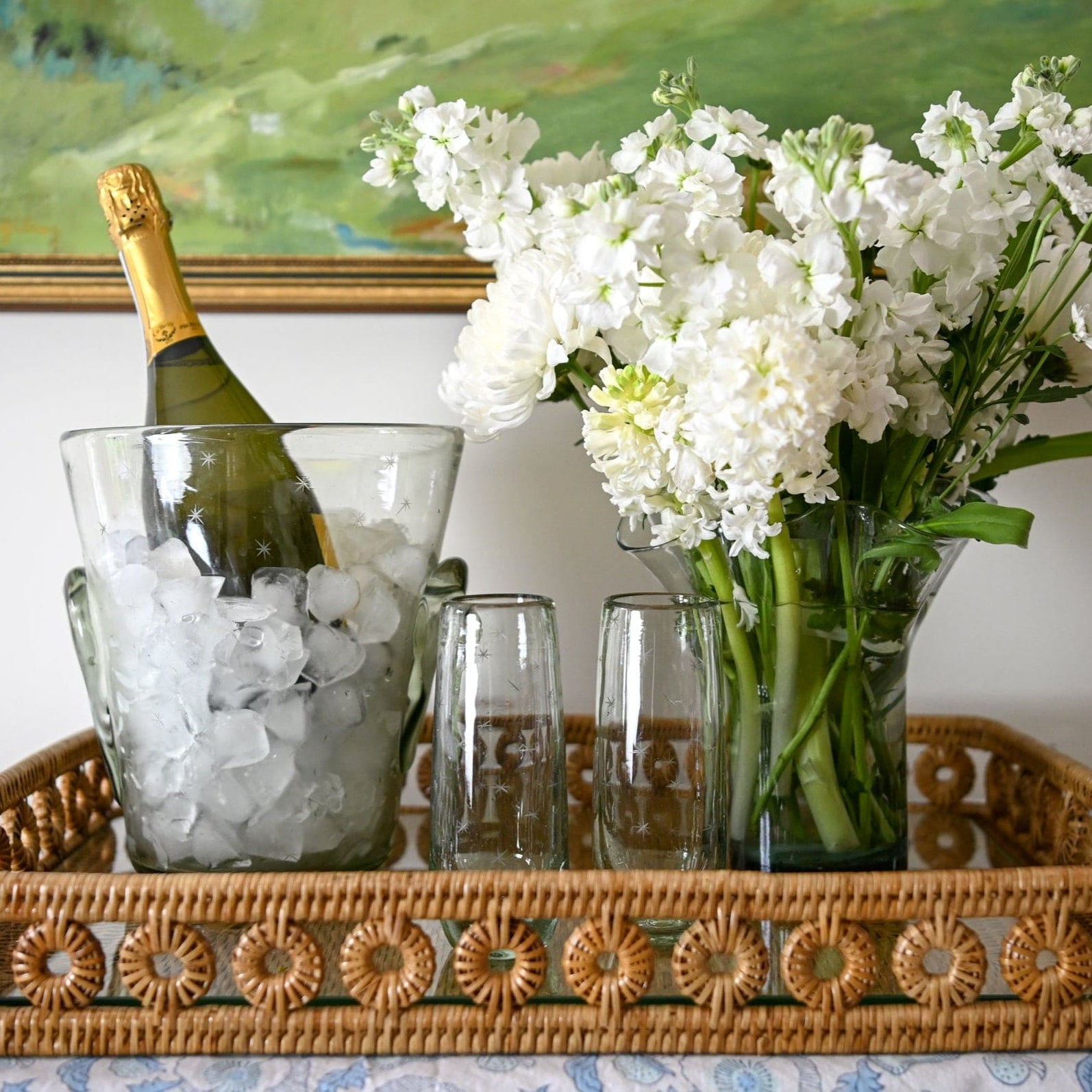 Bottle of champagne with ice, glasses, and a vase of white flowers on a decorative tray.